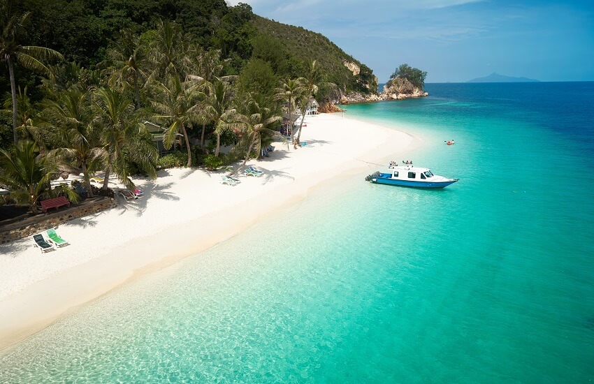 Powder-white beach on Rawa Island, Malaysia, with turquoise waters, palm-lined shore and a small boat floating offshore.