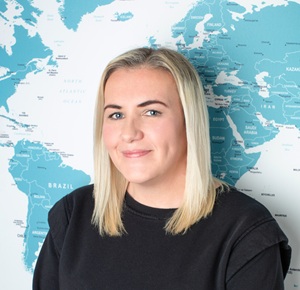 Profile photo of Faye standing in front of the office world map, smiling naturally.
