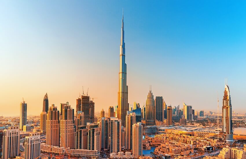 Burj Khalifa at sunrise, rising high above surrounding skyscrapers under a clear blue sky with warm morning light.