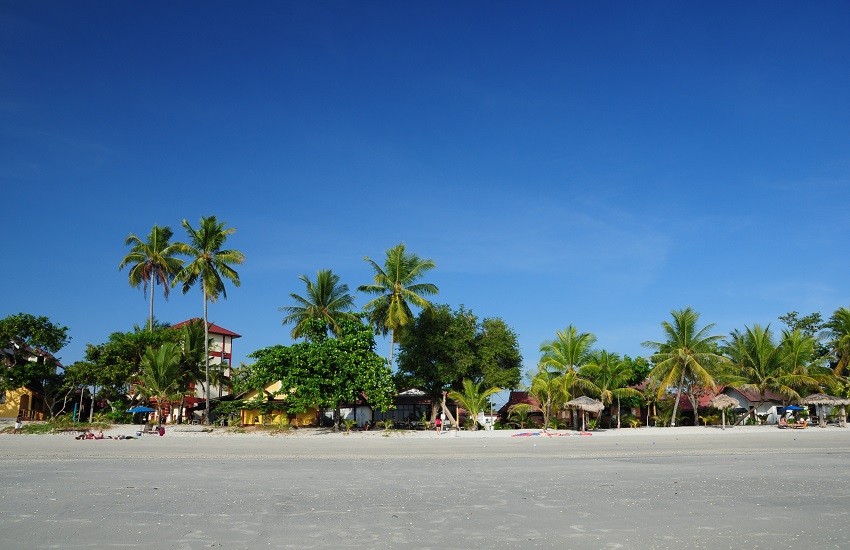 White-sand beach on Langkawi, with small beachside buildings dotted along the shoreline beneath a bright blue sky.