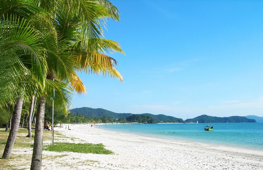 White-sand beach at Pantai Cenang on Langkawi, with turquoise waters and palm trees beneath a clear blue sky.
