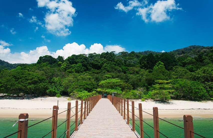 Kerachut Beach in Penang National Park, viewed from a pier leading towards the beach with lush greenery behind.