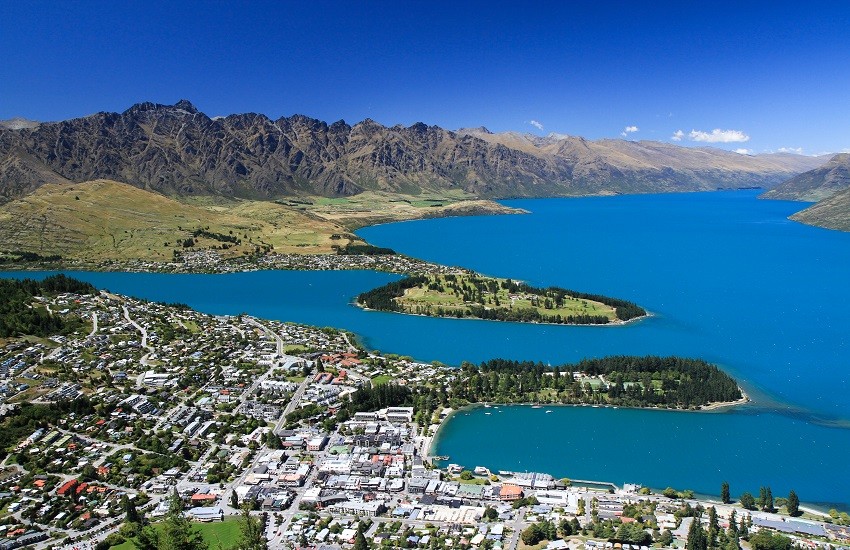 Aerial view of Queenstown in New Zealand with mountains rising behind the town and vivid blue water surrounding the city