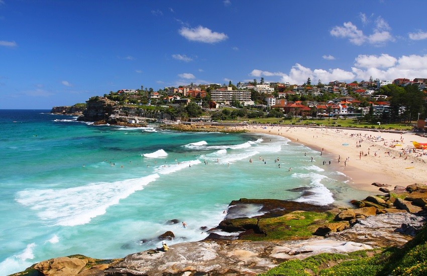 Bronte Beach in Sydney with roaring waves, people enjoying the sea and shoreline, and a clear sky with scattered clouds.