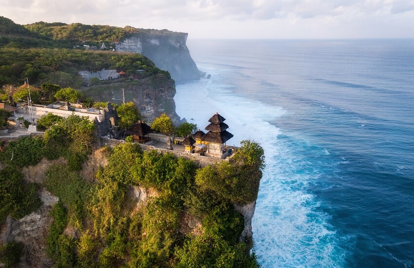 Uluwatu Temple perched on a cliff in Bali with waves crashing below and rugged cliffs stretching into the distance.