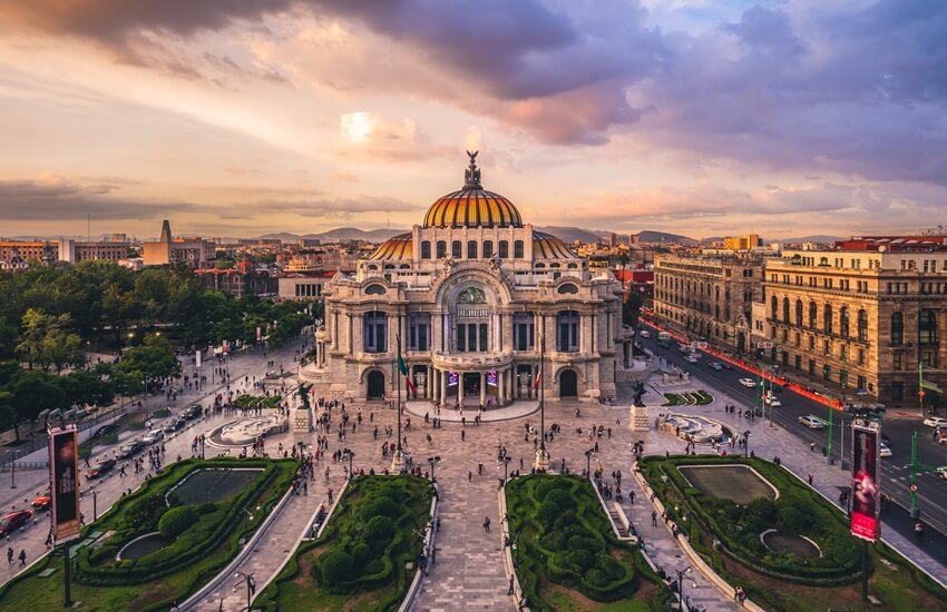 The Palacio de Bellas Artes in Mexico City at sunset with a pink sky and a few people scattered across the plaza in front.