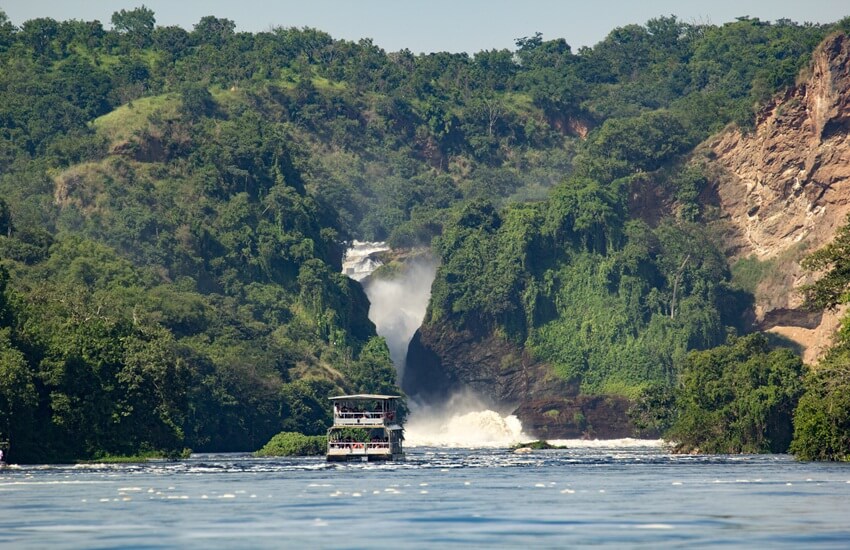 View from the waterline looking towards Murchison Falls in the centre, surrounded by rocky hills covered in trees and vegetation.