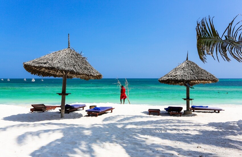 Turtle Beach in Watamu, Kenya, with loungers on white sand, turquoise waters in the background and a person in red garments standing at the water’s edge.