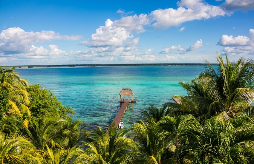 View of Lake Bacalar with turquoise water stretching into the distance, bright green palms in the foreground, a central pier and a small boat docked under a blue sky with scattered clouds.
