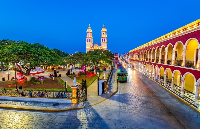 Campeche’s central square at twilight with trees in the plaza, surrounding buildings to the right, a cathedral in the background and a few people dotted around.