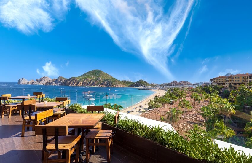 A rooftop restaurant in Los Cabos with beach and hotel resorts in the background under a bright blue sky with a swirl of cloud.
