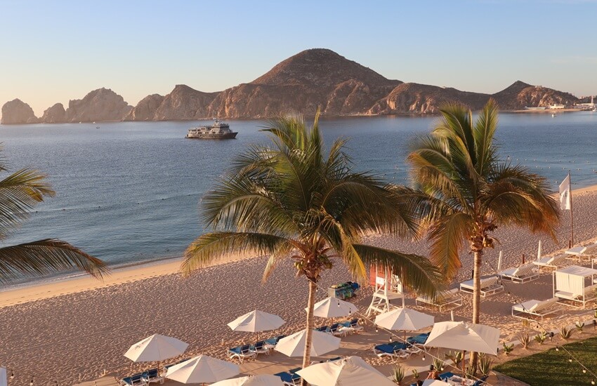 A quiet early-morning beach in Los Cabos with clear blue skies, mountain backdrops, and empty loungers outside a beachfront hotel.