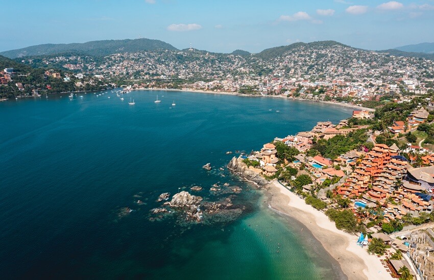 Aerial shot of Zihuatanejo’s beach with yachts in the bay, blue sea and sky, and houses and hotels scattered across the hillsides.
